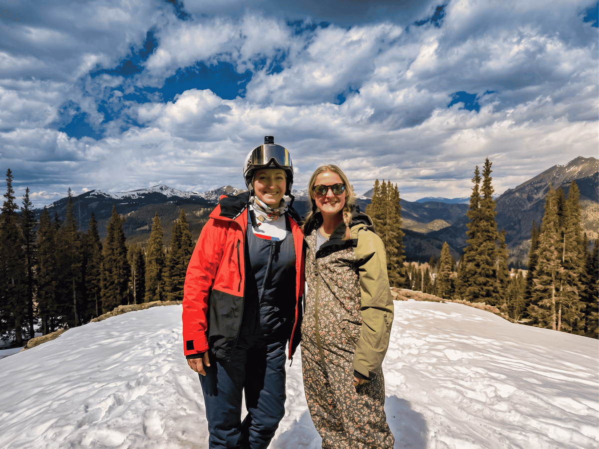 Two people standing on a snowy mountain with a scenic background of trees and mountains.